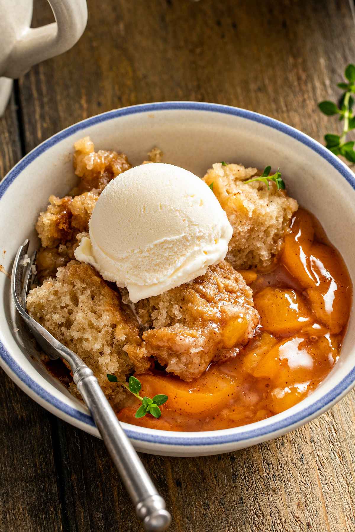 Close up of a bowl of crockpot peach cobbler topped with vanilla ice cream and garnished with thyme sprigs, with a spoon.