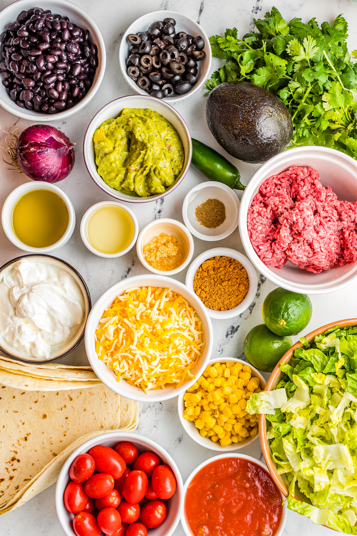 From top left: Black beans, sliced olives, cilantro, avocado, red onion, jalapeno, lime, olive oil, lime juice, garlic, salt and pepper, ground beef, taco seasoning, sour cream, shredded cheese, corn, shredded lettuce, tortillas, grape tomatoes, and salsa.