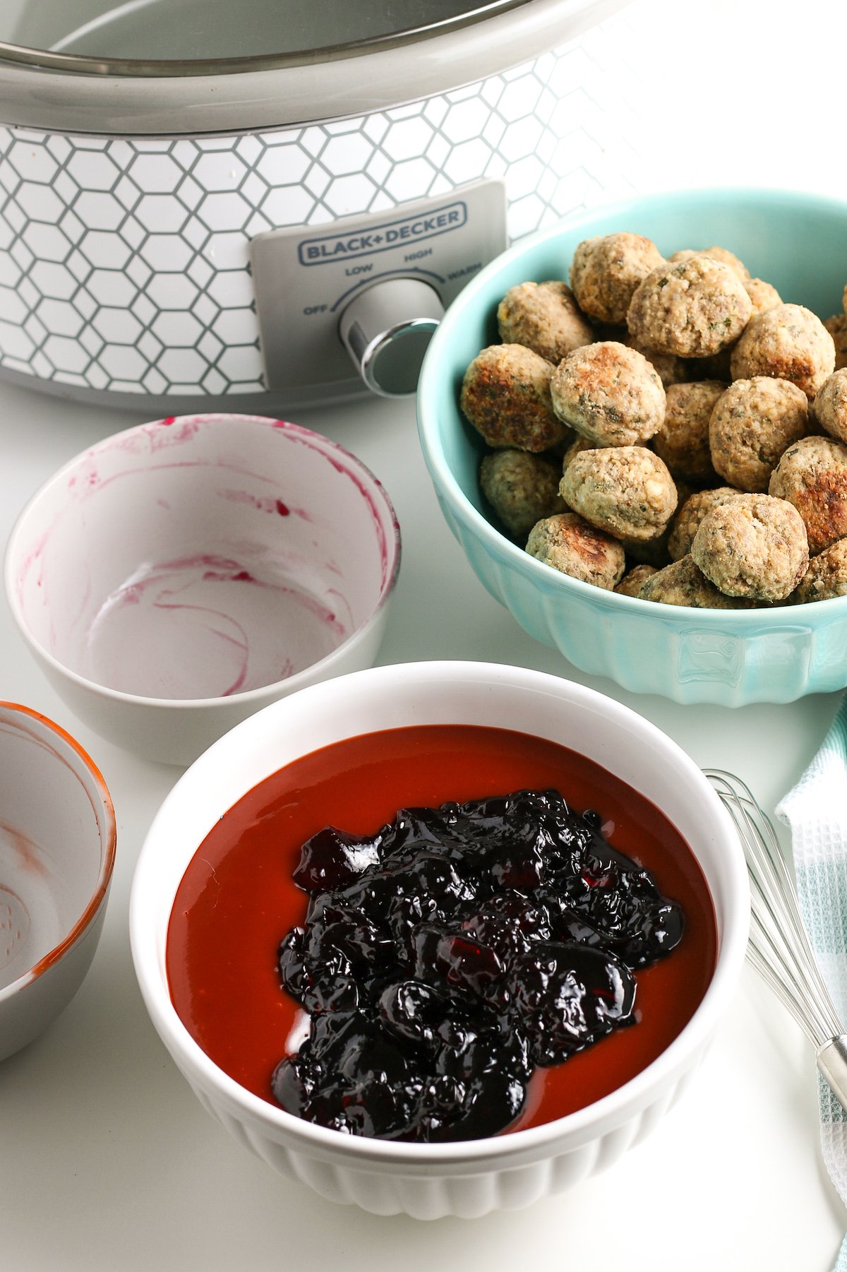 Jelly and barbecue sauce in a bowl, next to a dish of meatballs and a crockpot.