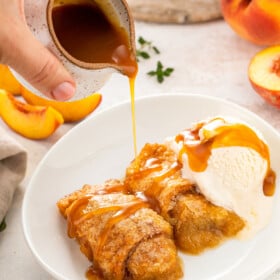Peach dumplings in a white bowl with caramel sauce being poured on top.