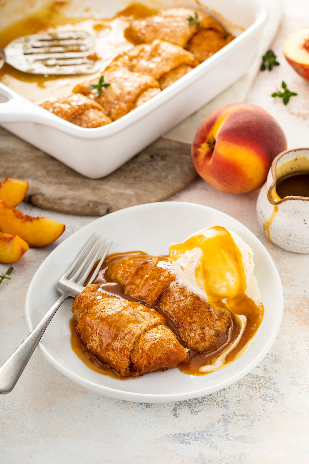 A pan of peach dumplings, with a small dessert plate served with dumplings, ice cream, and caramel sauce. A fresh peach and small pitcher of sauce are near the plate on the table.
