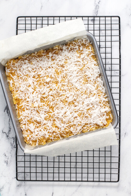 A square, parchment-lined pan of bar cookies topped with fresh coconut.