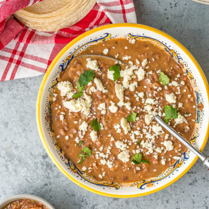 A large bowl of refried beans, garnished with cheese crumbles and fresh cilantro, next to a stack of tortillas in a cloth napkin.