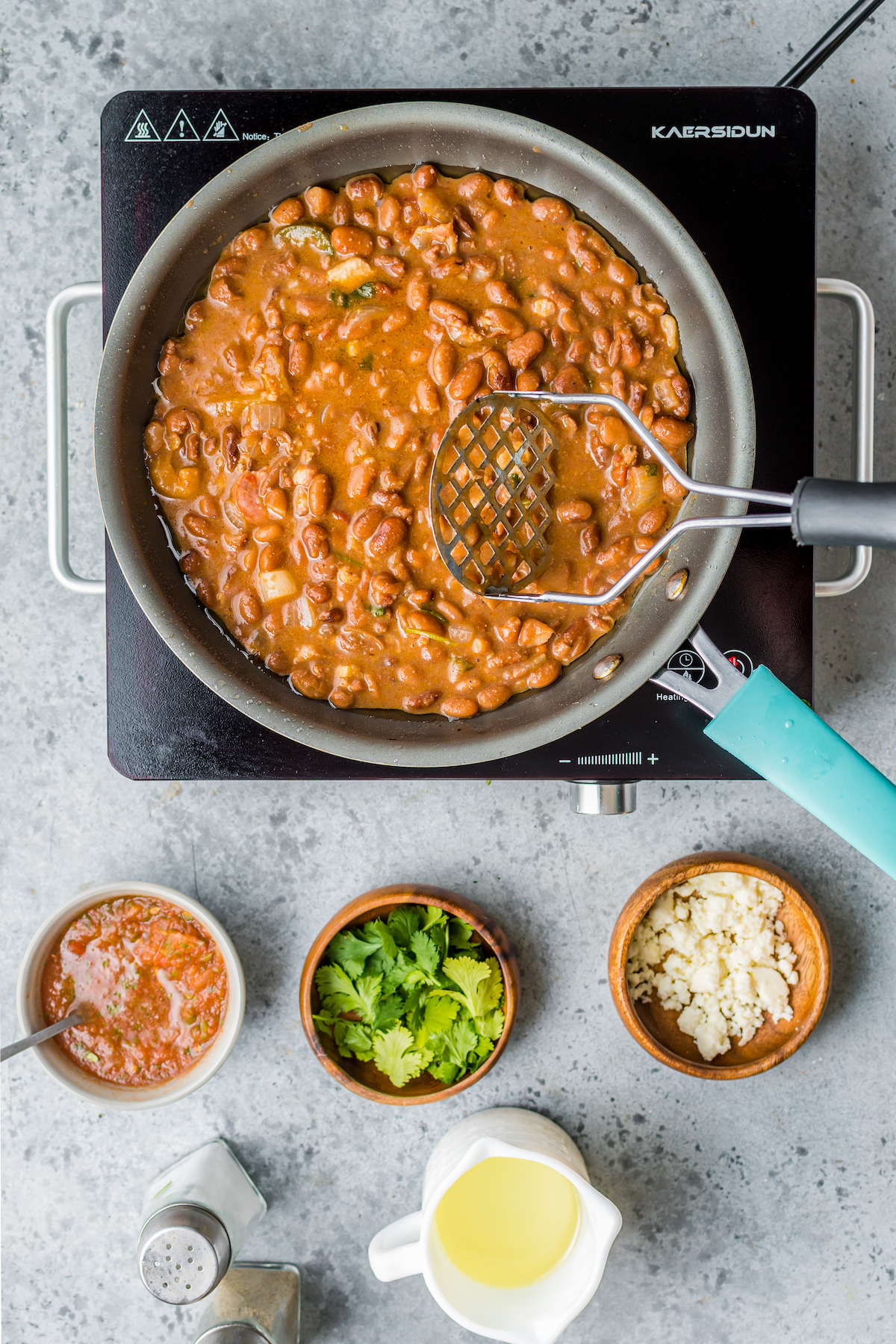 Beans cooking in a skillet. A potato masher is resting in the beans, and small dishes of salsa, cilantro, and queso fresco are nearby.