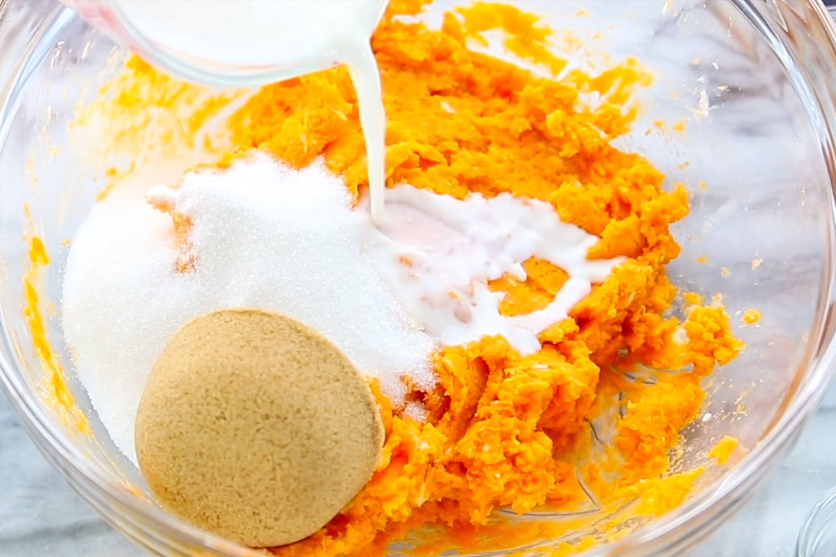 Mashed sweet potatoes in a bowl with brown sugar, granulated sugar and milk being poured in.