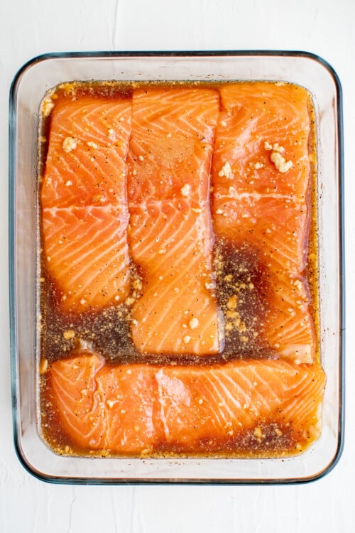 Salmon fillets lined up in a small rectangular glass pan, with marinade poured over them.