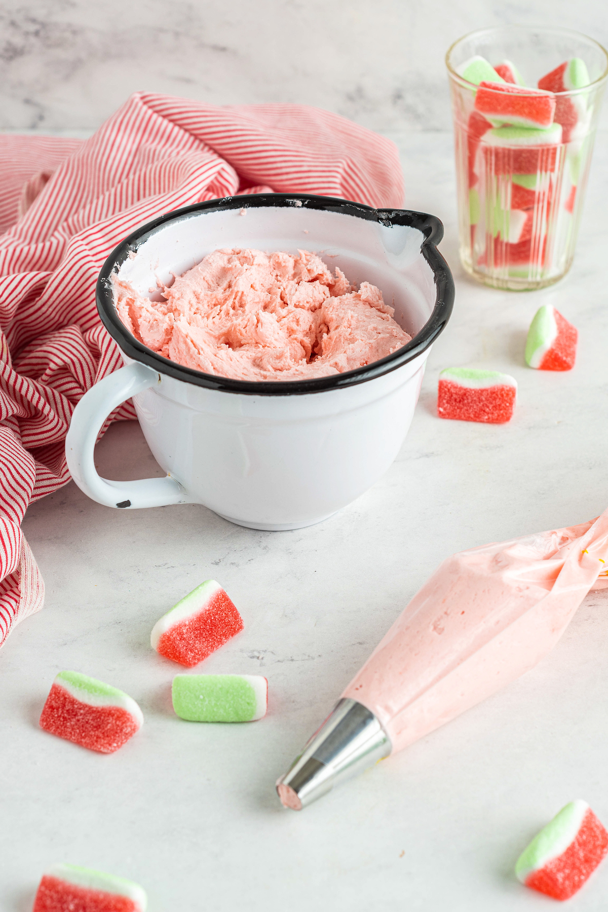 A large measuring cup filled with pink watermelon buttercream, next to a piping bag and scattered watermelon candies.