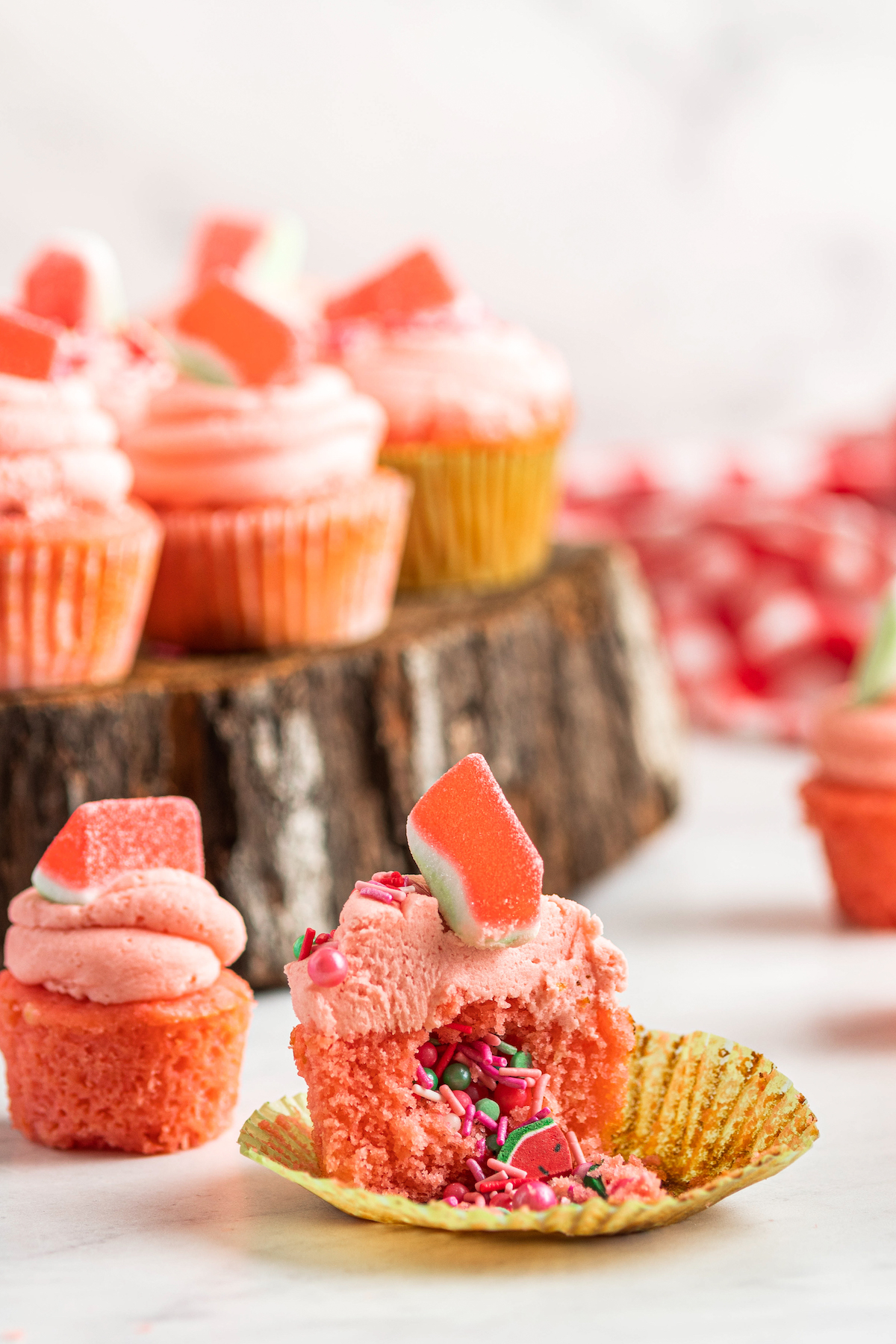 Pink watermelon cupcakes arranged around a decorative slab of wood, with one cupcake cut in half to reveal the surprise sprinkle center.