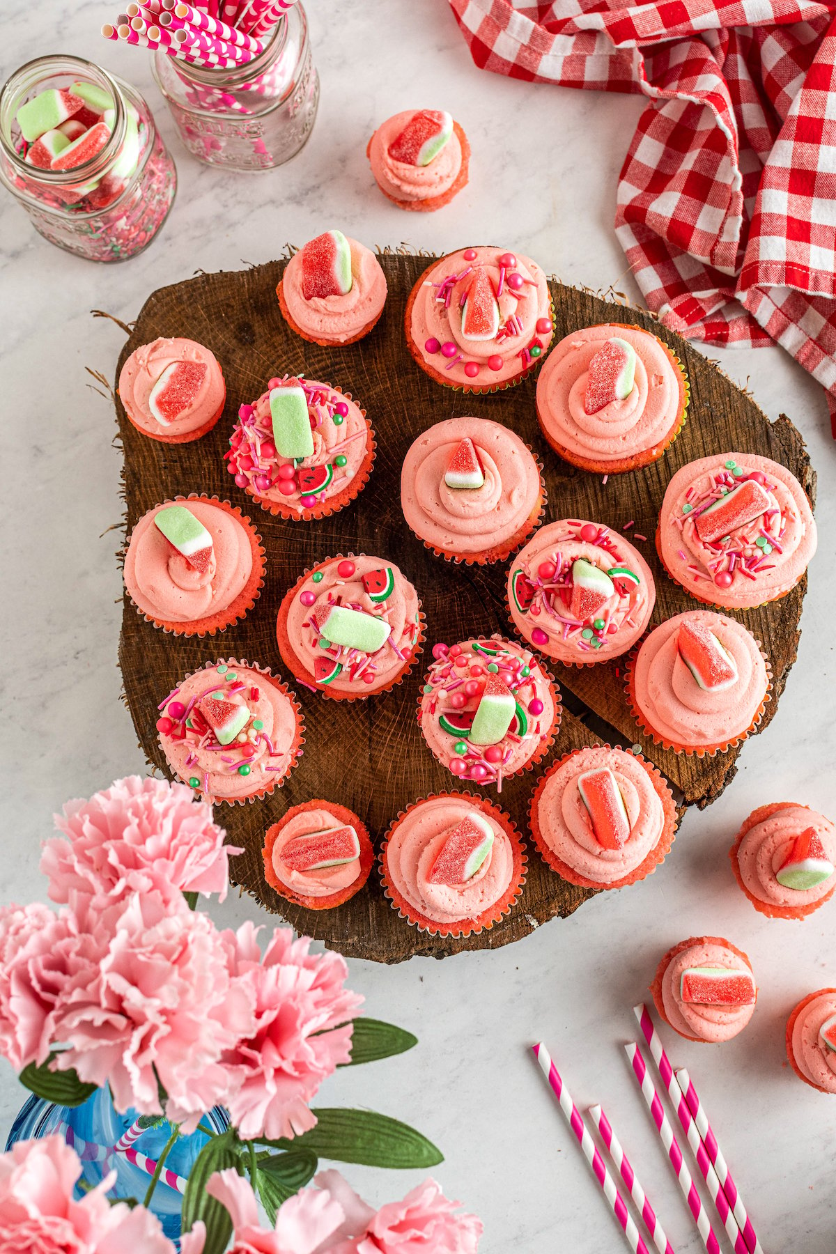 Top view of decorated watermelon cupcakes on a wooden platter, next to scattered cupcakes and pink flowers.