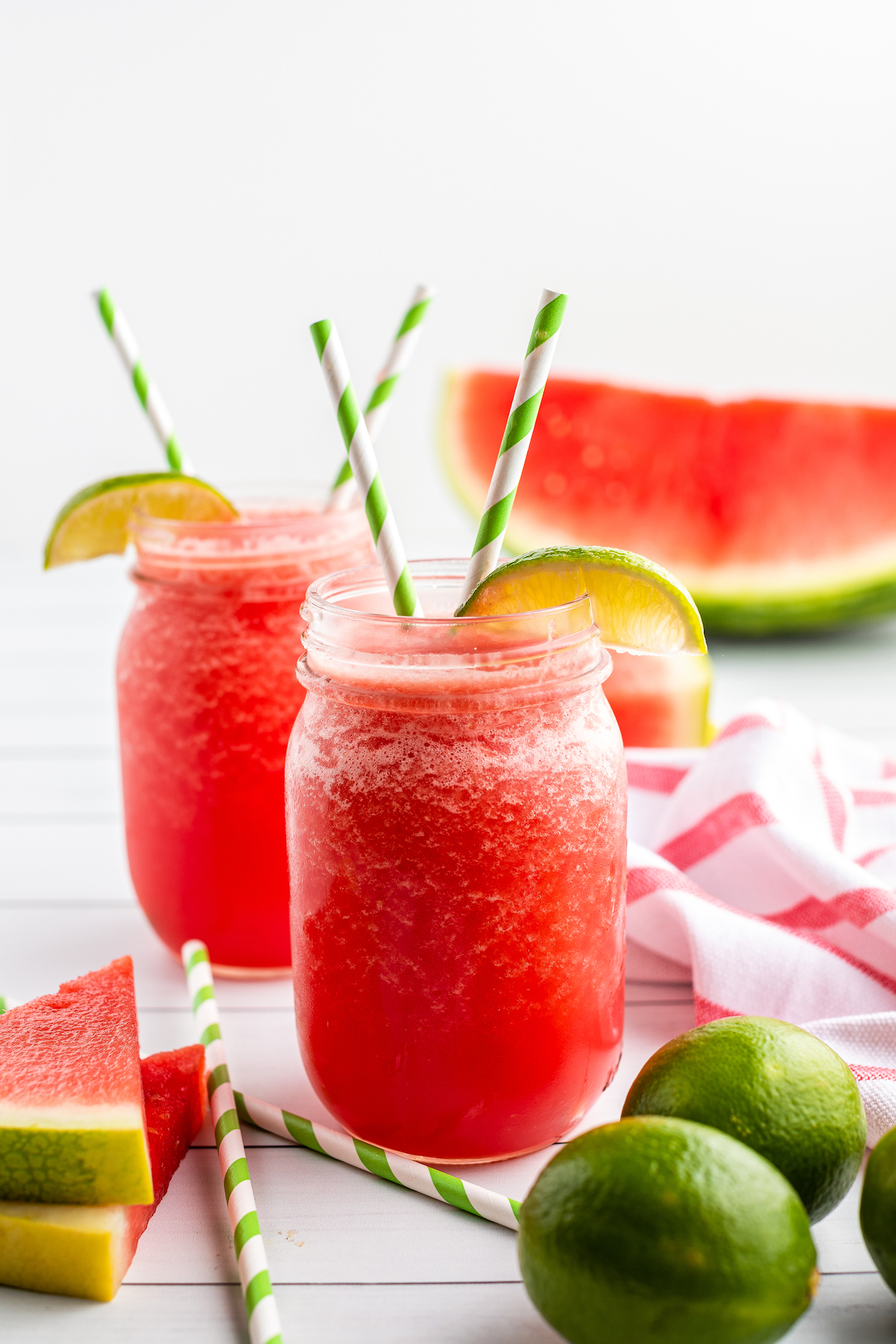 Two watermelon limeade slushies in mason jars, side-by-side, garnished with lime wedges and striped straws.