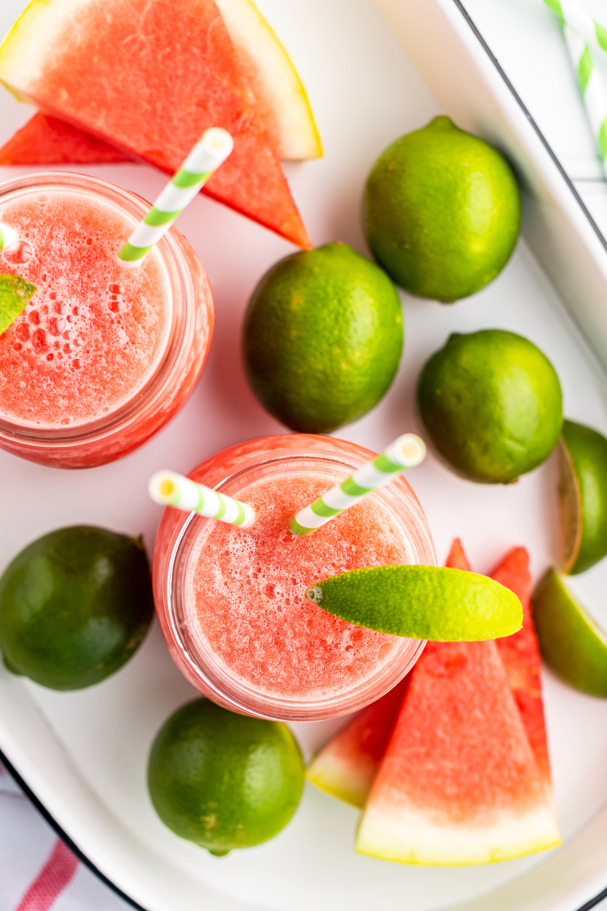Top view of two watermelon limeade slushies in mason jars side-by-side, garnished with lime wedges and striped straws, on a tray with watermelon slices and limes.