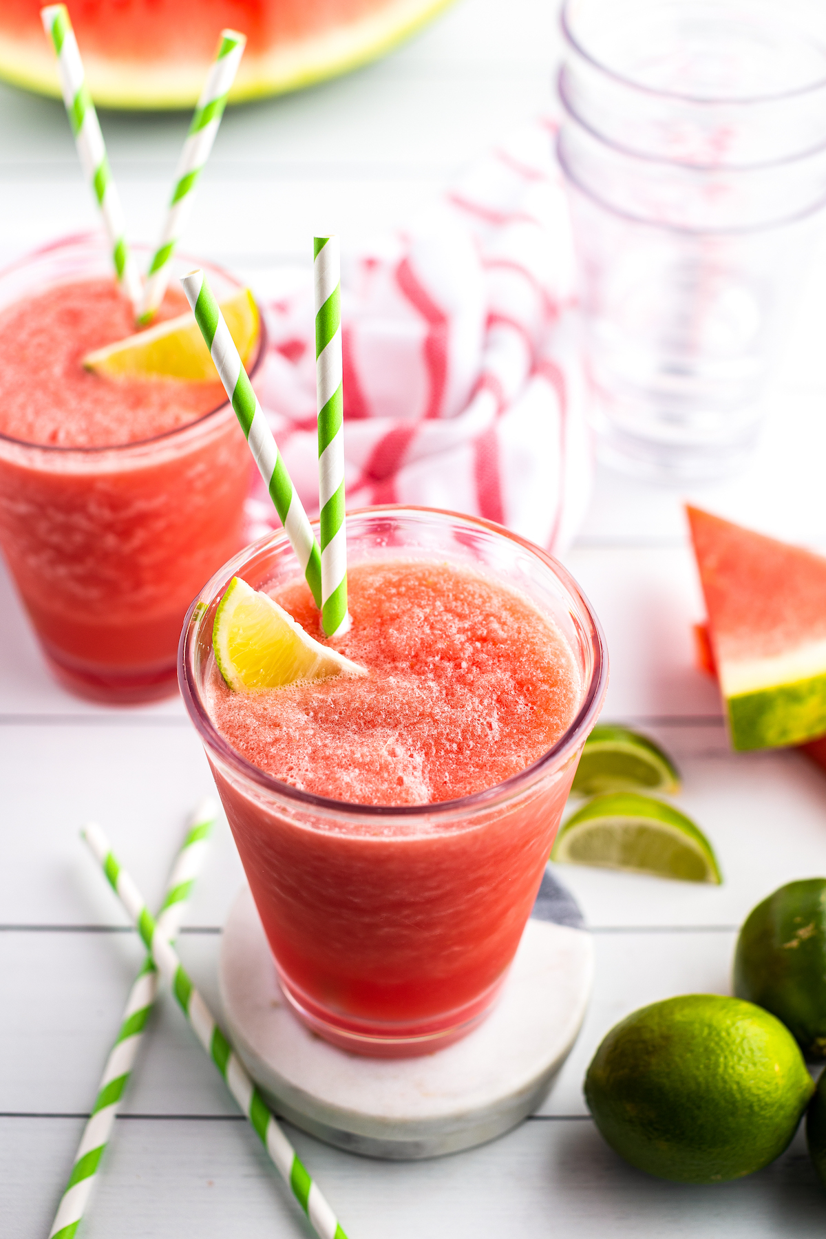 Two side-by-side watermelon limeade slushies in glasses garnished with lime wedges and striped straws.