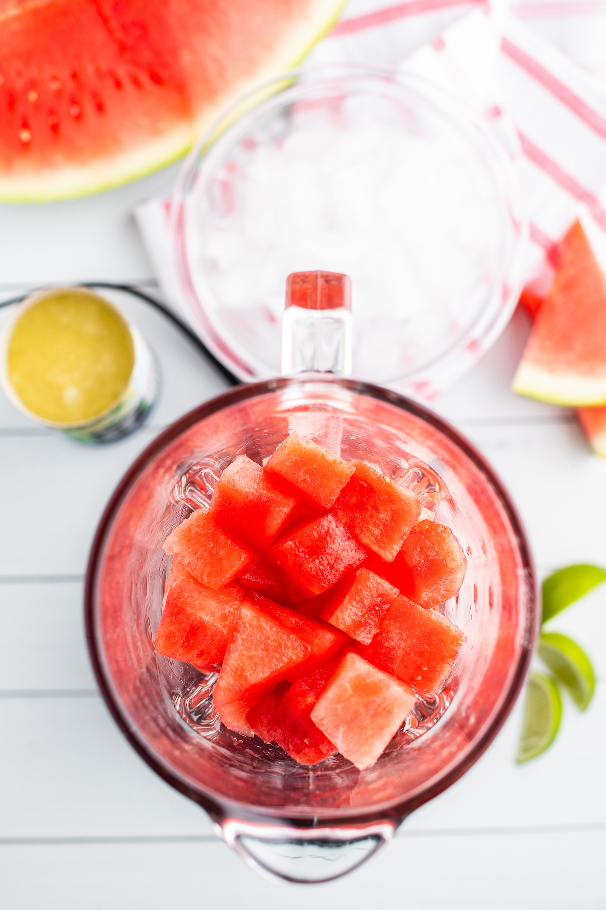Top view of watermelon cubes in a blender next to a half a watermelon, a bowl of ice and a can of frozen limeade.
