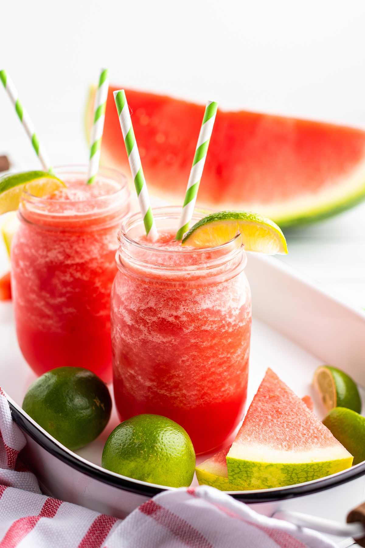 Two watermelon limeade slushies in mason jars side-by-side, garnished with lime wedges and striped straws, on a tray with watermelon slices and limes.