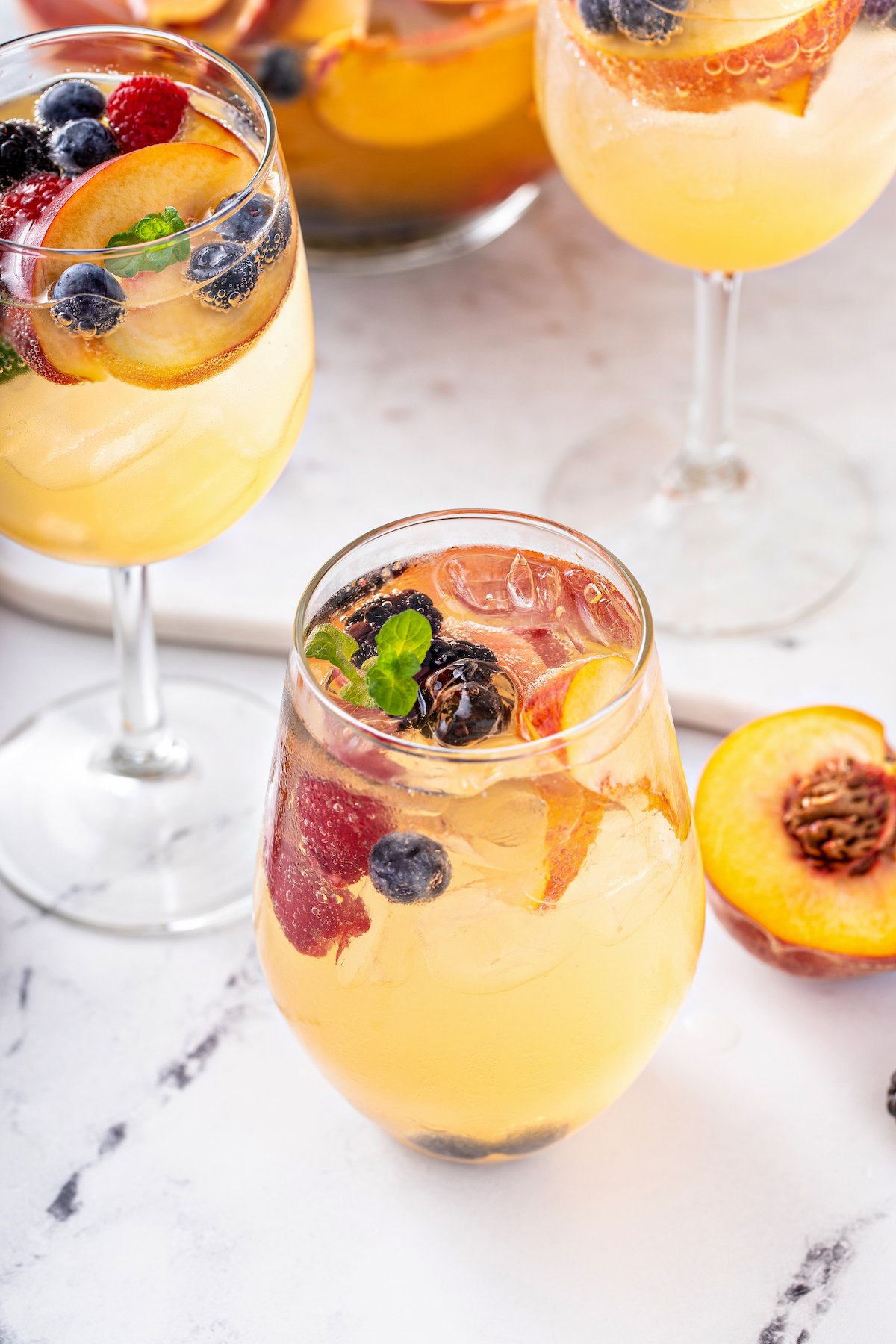 A glass of white wine cocktail, garnished with fruit and mint. Other glasses and half a peach are visible in the background.