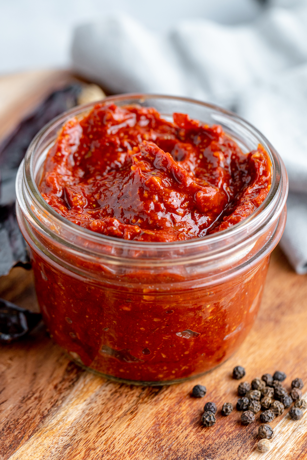 A small jar of thick red sauce on a wooden work surface, with a cloth napkin and peppercorns in the background.