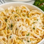 A bowl of creamy fettuccine Alfredo pasta made with rich Alfredo Sauce With Cream Cheese, garnished with chopped parsley and black pepper, with a plate of pasta and fresh herbs in the background.