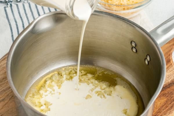 Cream is being poured into a saucepan containing melted butter and sautéed garlic, with shredded cheese in a bowl and a striped towel visible in the background.