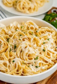 A bowl of creamy fettuccine Alfredo pasta made with rich Alfredo Sauce With Cream Cheese, garnished with chopped parsley and black pepper, with a plate of pasta and fresh herbs in the background.