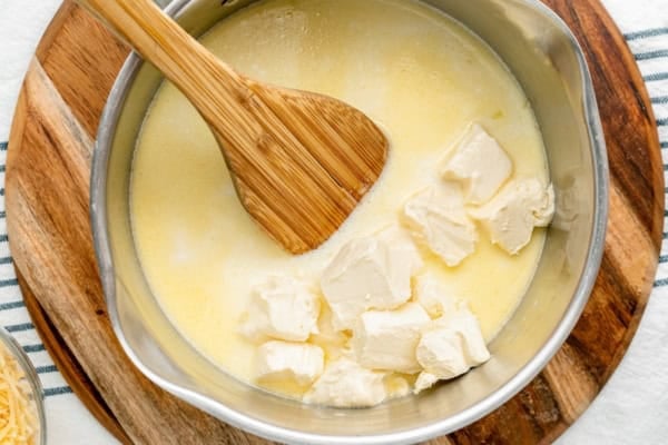 A wooden spoon stirs cream cheese into melted butter in a metal saucepan, sitting on a wooden board. The mixture appears to be in the process of melting and combining.