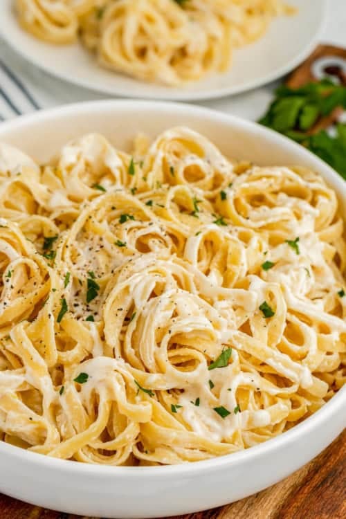 A bowl of creamy fettuccine Alfredo pasta made with rich Alfredo Sauce With Cream Cheese, garnished with chopped parsley and black pepper, with a plate of pasta and fresh herbs in the background.