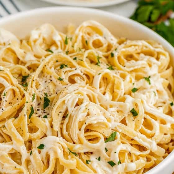 A bowl of creamy fettuccine Alfredo pasta made with rich Alfredo Sauce With Cream Cheese, garnished with chopped parsley and black pepper, with a plate of pasta and fresh herbs in the background.