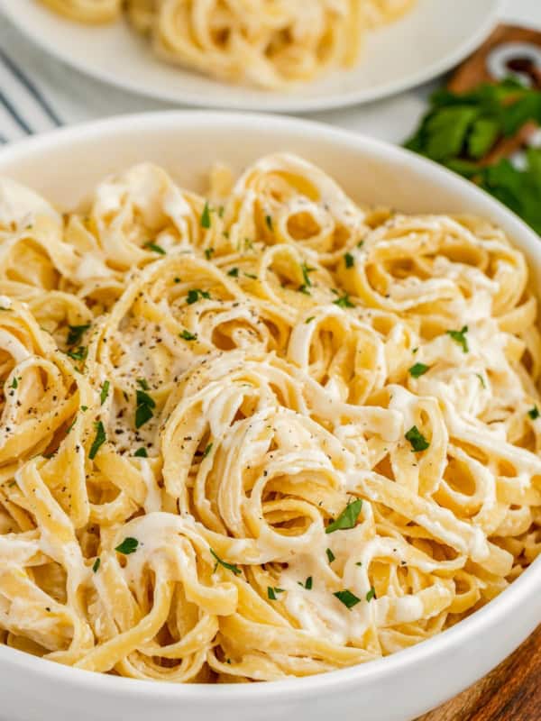 A bowl of creamy fettuccine Alfredo pasta made with rich Alfredo Sauce With Cream Cheese, garnished with chopped parsley and black pepper, with a plate of pasta and fresh herbs in the background.