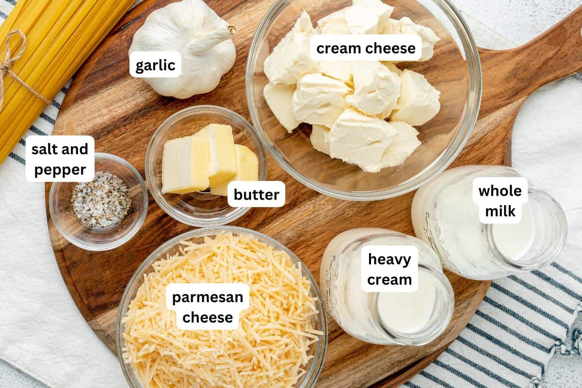 Alfredo sauce with cream cheese makes for a garlicky, cheesy pasta dinner that everyone will love! Fool proof and ready in 10 minutes. Labeled ingredients for a pasta dish on a wooden board: uncooked spaghetti, whole garlic, cream cheese, butter, heavy cream, whole milk, grated parmesan cheese, and a small bowl of salt and pepper—everything you need to create a rich Alfredo Sauce With Cream Cheese.