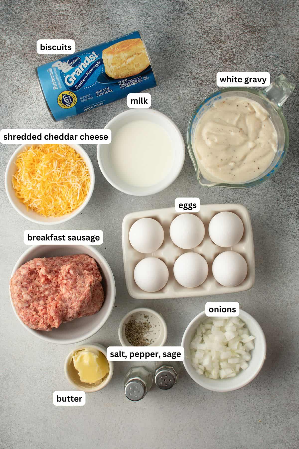 The ingredients for biscuits and gravy casserole recipe in order from top to bottom: biscuits, shredded cheddar cheese, milk, white gravy, breakfast sausage, eggs, onions, salt, pepper, sage, and butter.