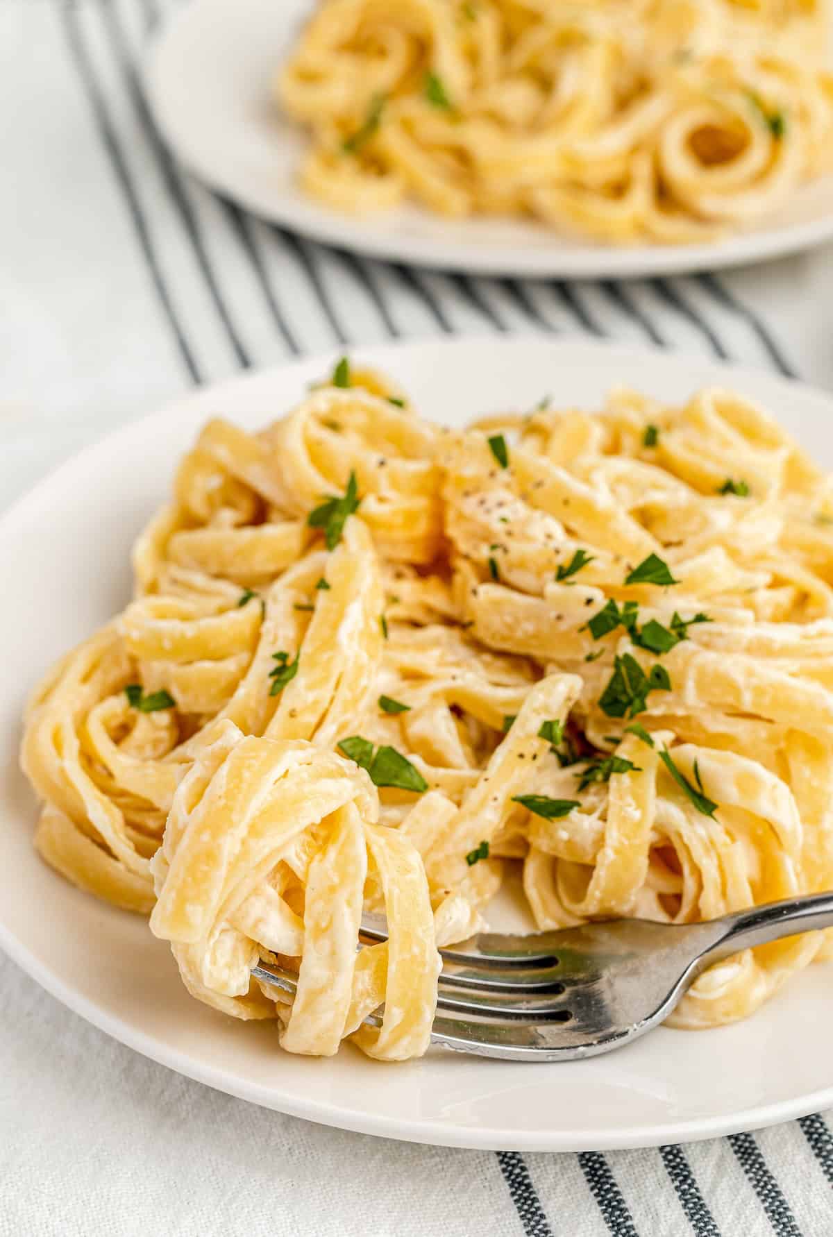 Alfredo sauce with cream cheese makes for a garlicky, cheesy pasta dinner that everyone will love! Fool proof and ready in 10 minutes. A plate of creamy fettuccine alfredo made with a luscious Alfredo Sauce With Cream Cheese, garnished with chopped parsley. A fork twirls some noodles in the foreground on a white plate, with another plate of pasta blurred in the background.