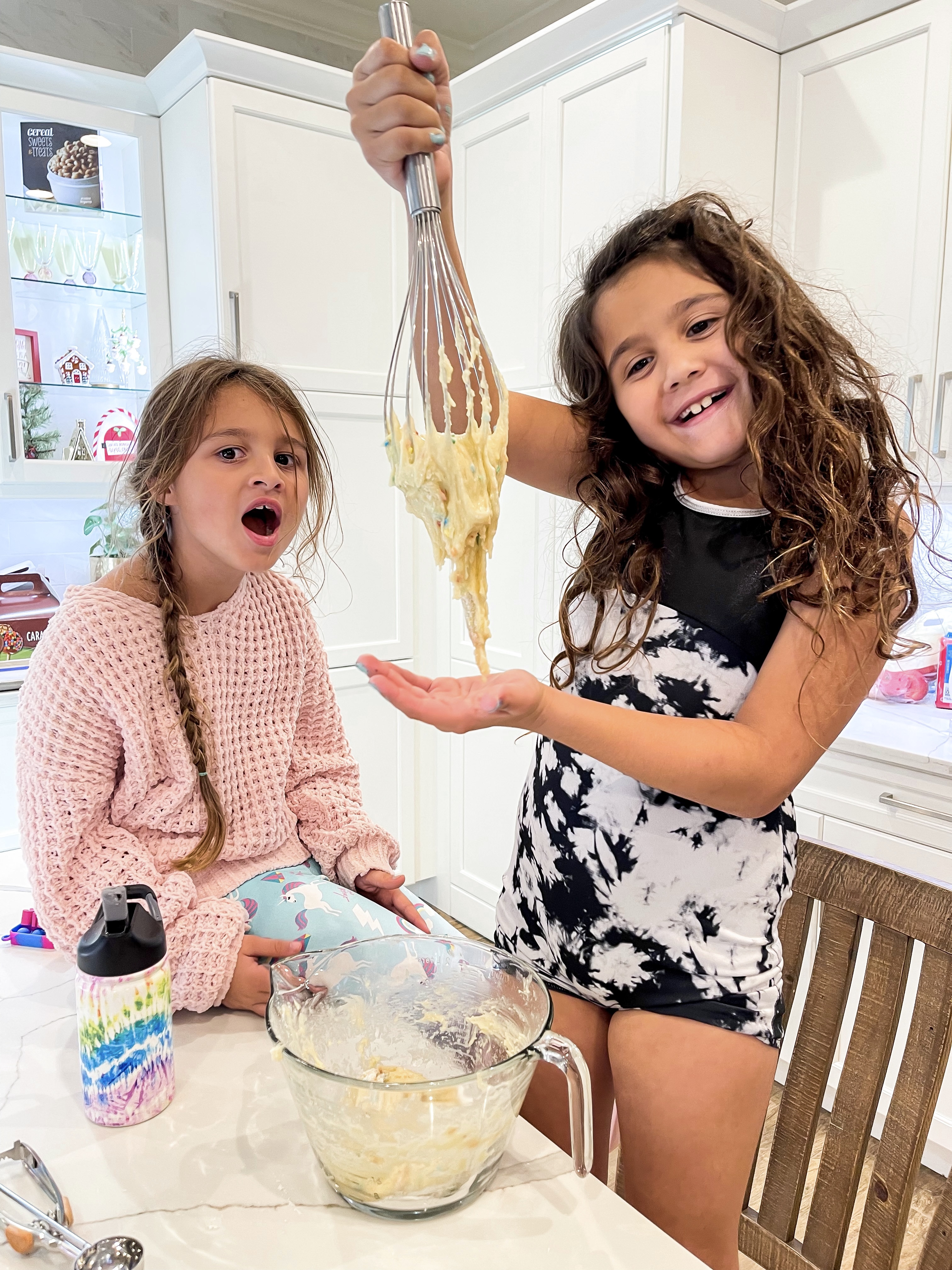 Two girls baking cookies with one girl holding a whisk.