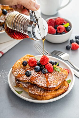 A stack of French toast pieces, topped with berries and powdered sugar. Maple syrup is being poured on top.