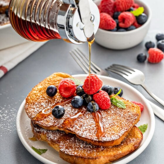 A stack of French toast pieces, topped with berries and powdered sugar. Maple syrup is being poured on top.