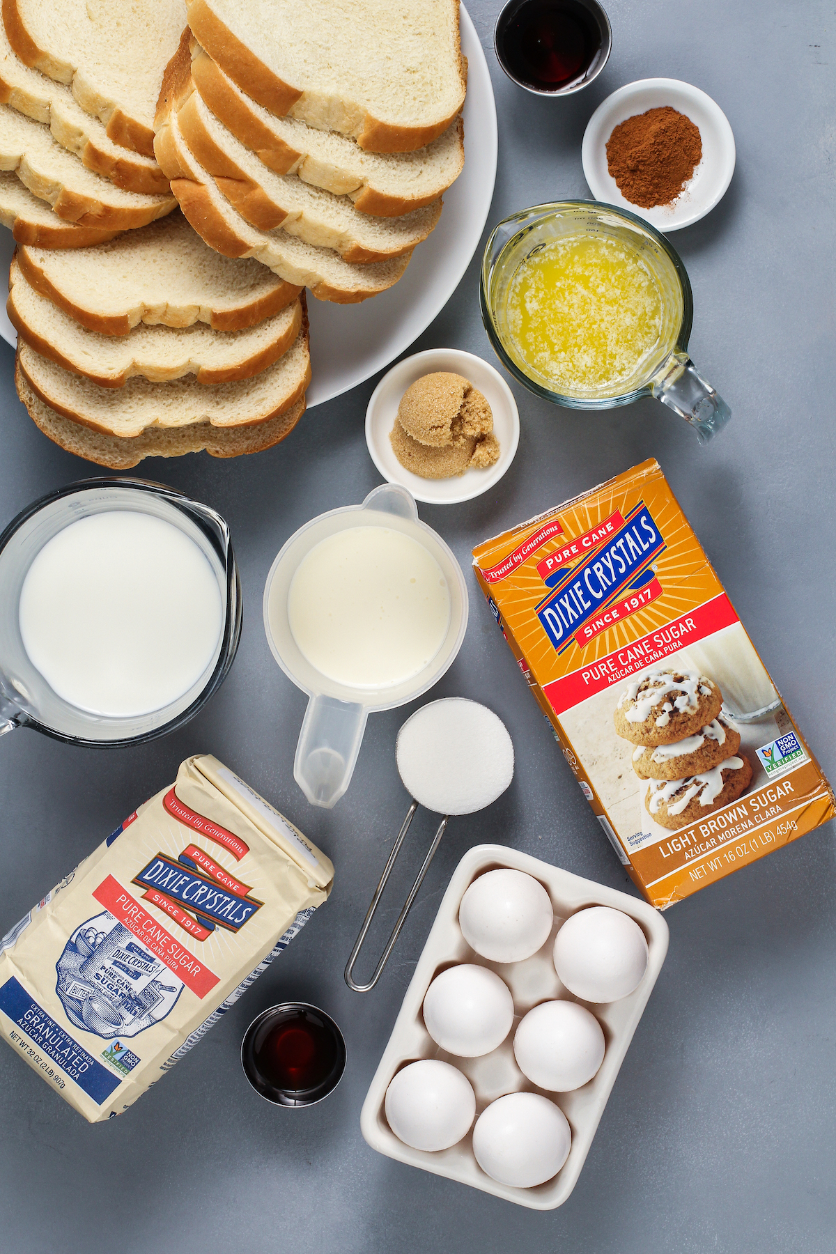 From top left: Sliced bread, vanilla, melted butter, cream, milk, cinnamon, granulated sugar, brown sugar, eggs.