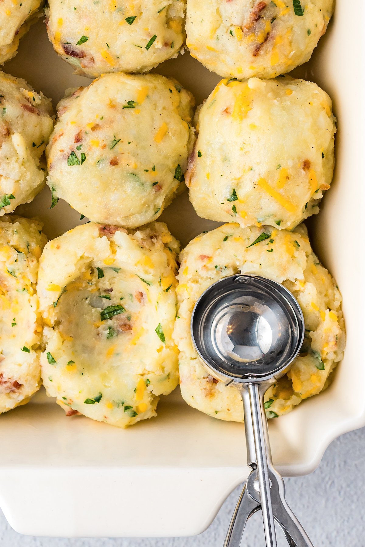 Mounds of mashed potatoes in a casserole dish with a cookie scoop pressing into the potatoes to make an indent.
