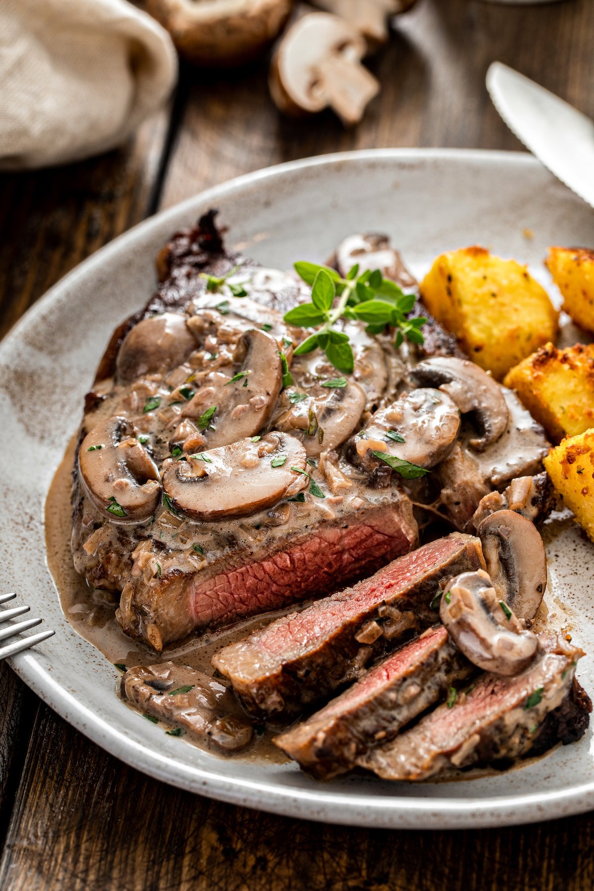 A sliced steak on a plate with mushroom sauce and roasted potatoes.