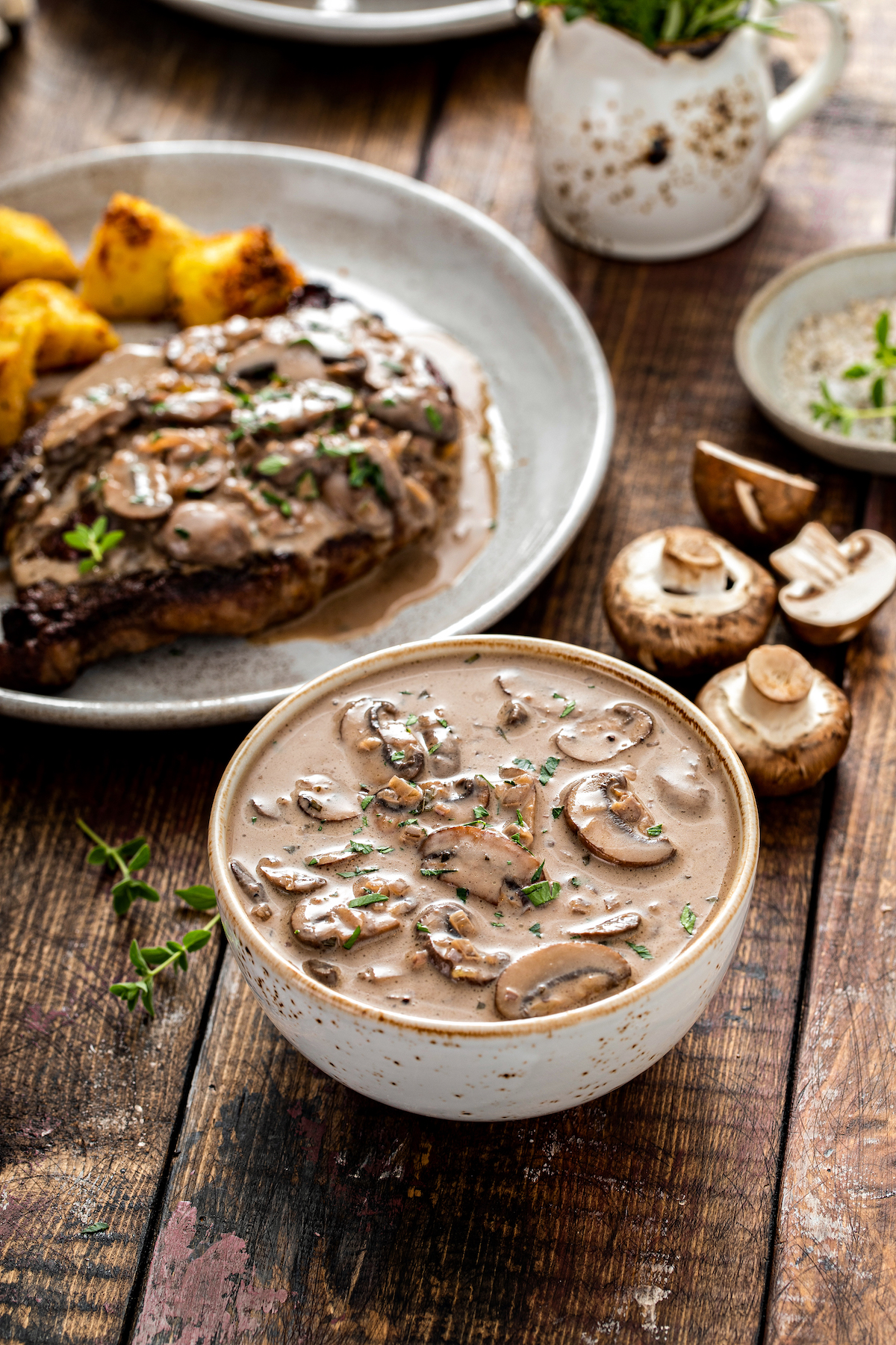 A bowl of creamy sauce next to a plate of steak and potatoes.