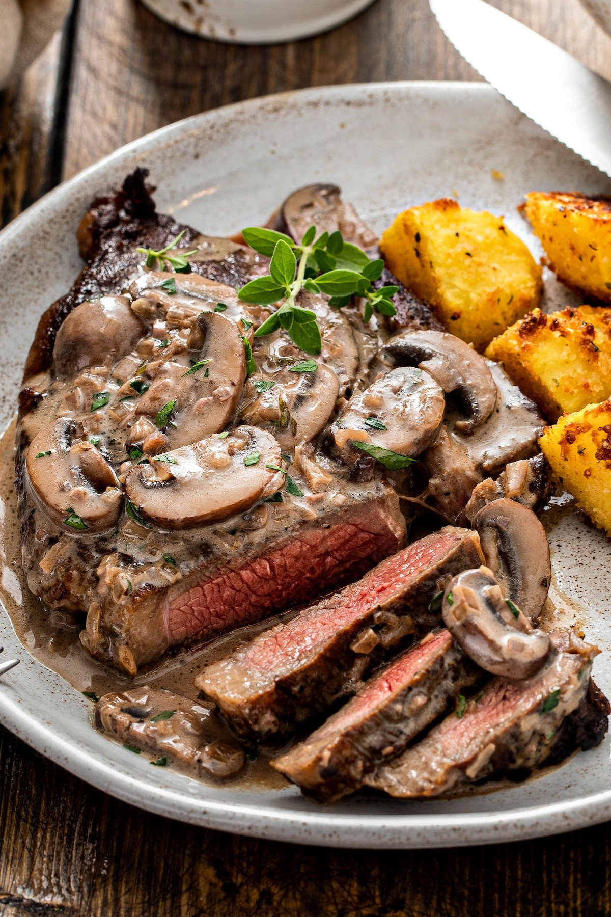 A platter of sliced, medium-well steak topped with creamy mushrooms and a garnish of parsley.