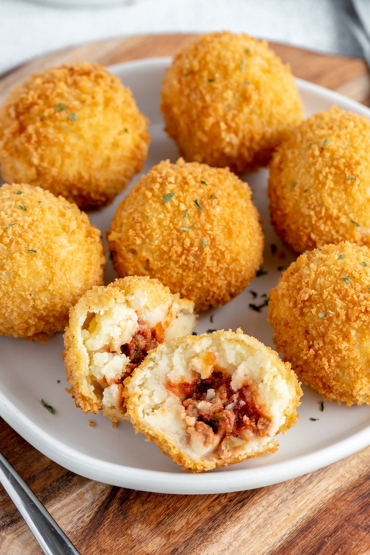 Seven fried mashed potato balls on a plate. One has been cut in half to show the beef filling.