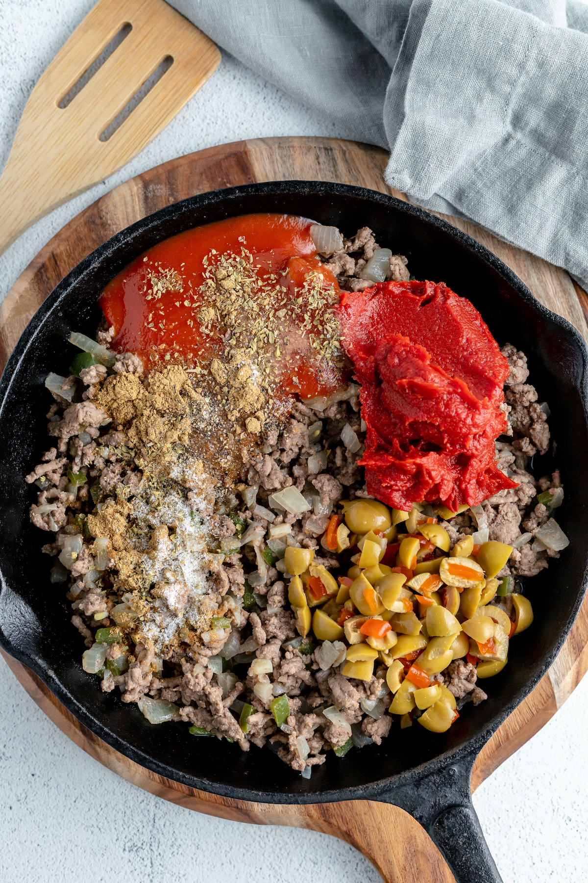 Ground beef, tomato paste, green olives, and other ingredients in a cast iron skillet.