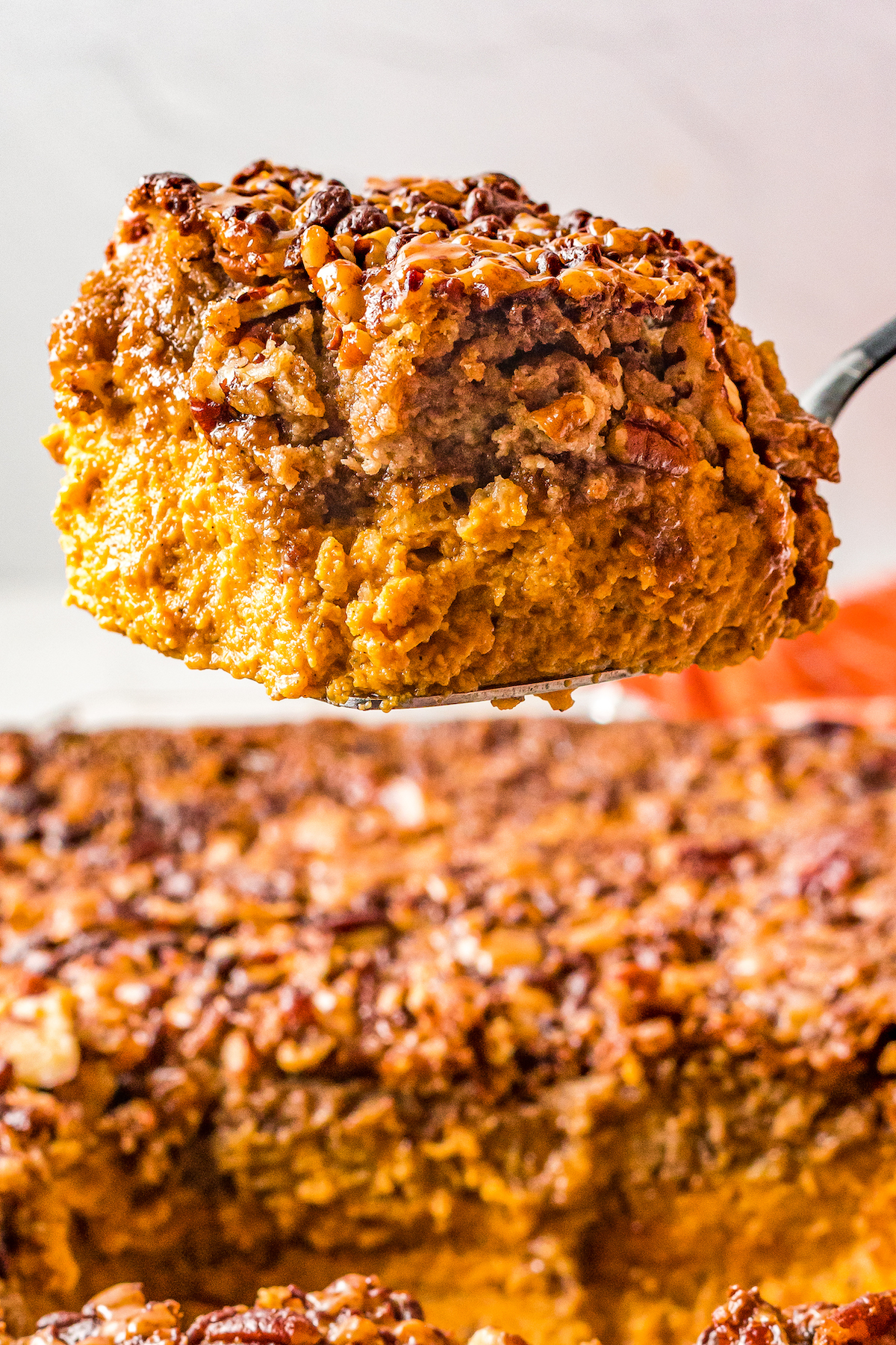 Close-up shot of a square of pumpkin dessert being held up to the camera on a cake server. The layers of pumpkin and crunchy topping are clearly defined.
