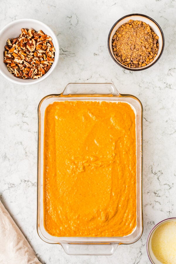 A rectangular glass baking dish filled with pumpkin batter, next to dishes of toppings and a bag of cake mix.