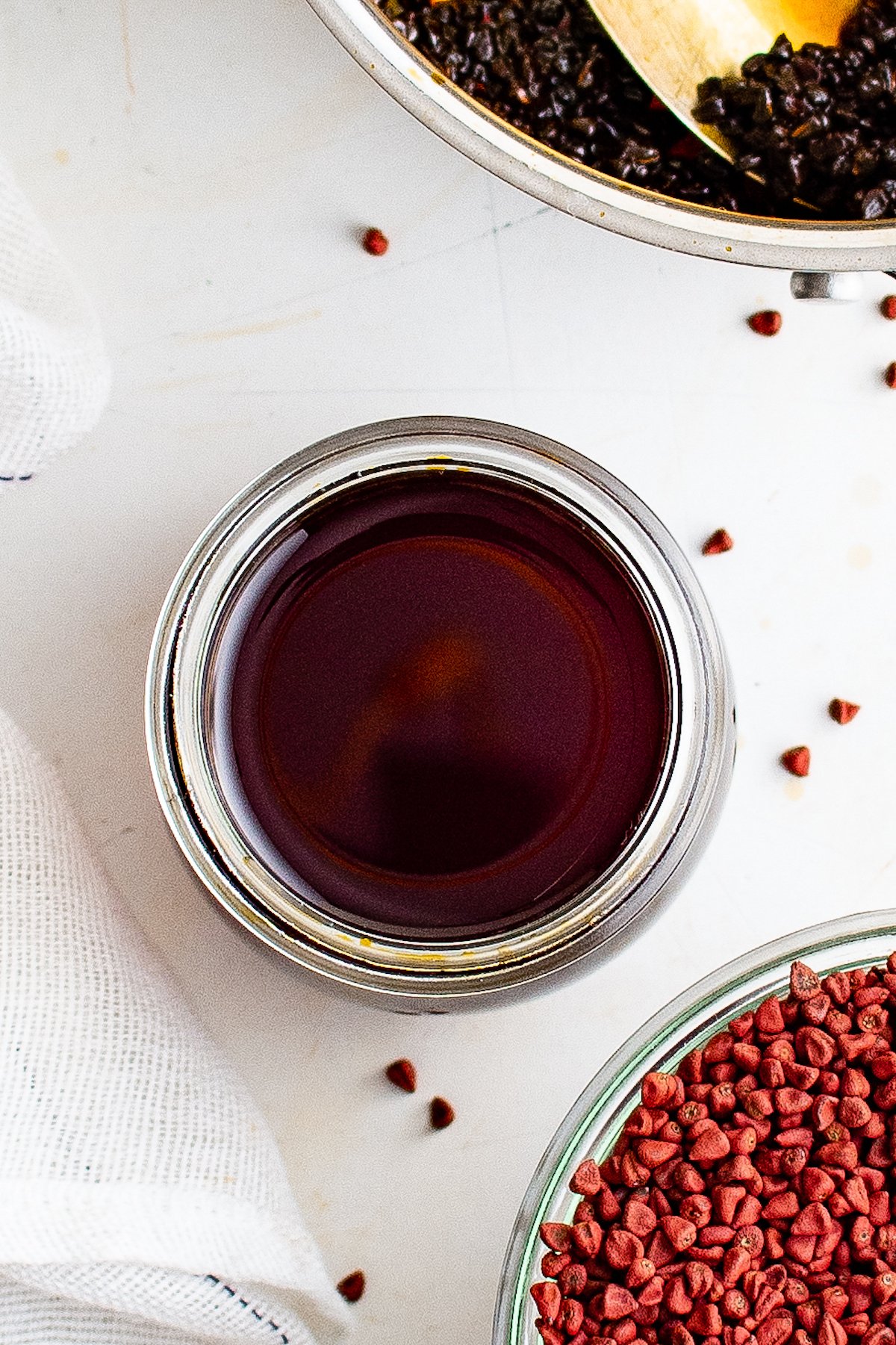 Overhead view of annatto oil in a glass jar, near a saucepan of leftover annatto seeds and a bowl of dried annatto seeds.