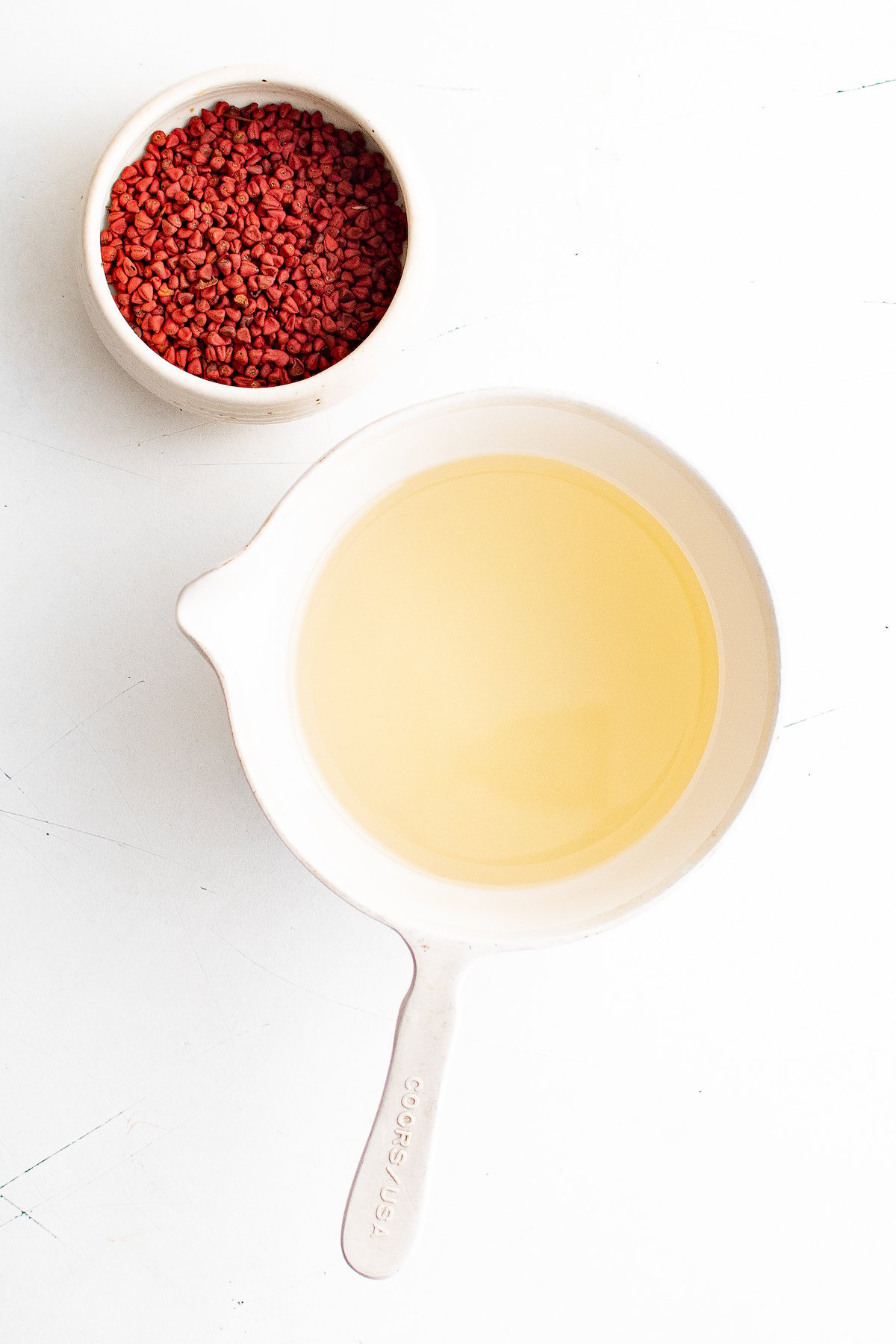 A dish of annatto seeds next to a measuring cup with oil in it.