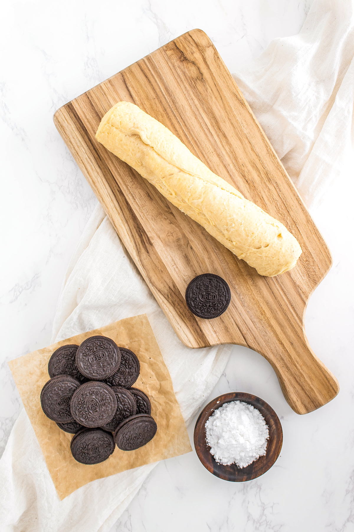a cutting board with a roll of crescent roll and one Oreo next to another pile of Oreos