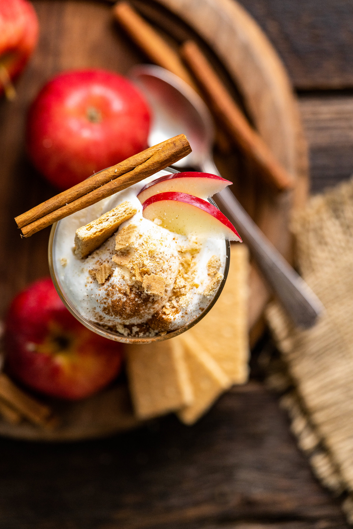 top view of a cocktail glass with apple cider and topped with whipped cream, apple slices, cinnamon, a piece of graham cracker and cinnamon stick alongside apples, graham crackers, and cinnamon sticks on a wooden platter