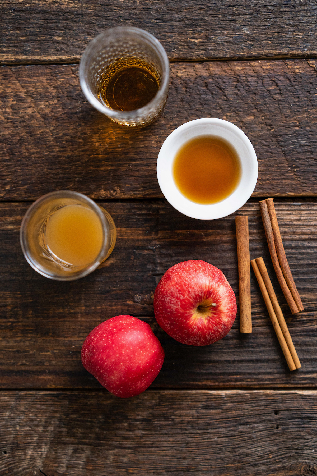 two apples, a glass with apple cider, small bowl with maple syrup and shot of rum on a wooden countertop