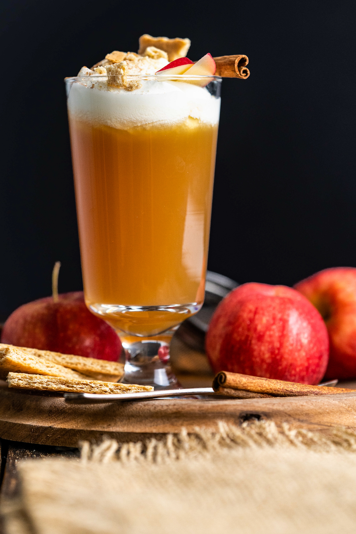 cocktail glass with apple cider and topped with whipped cream, apple slices, cinnamon, a piece of graham cracker and cinnamon stick alongside apples, graham crackers, and cinnamon sticks on a wooden platter