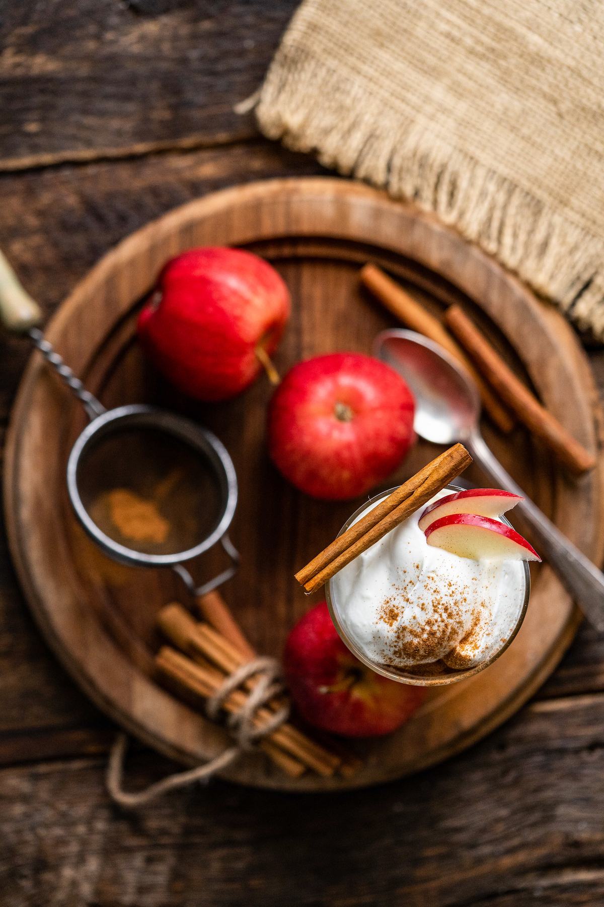 a cocktail glass with apple cider and topped with whipped cream, apple slices, cinnamon, a piece of graham cracker and cinnamon stick alongside apples, graham crackers, and cinnamon sticks on a wooden platter