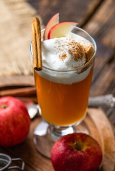 cocktail glass with apple cider and topped with whipped cream, apple slices, cinnamon, a piece of graham cracker and cinnamon stick